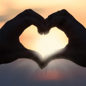 hands, fingers making the shape of a heart in foreground, background of sunset